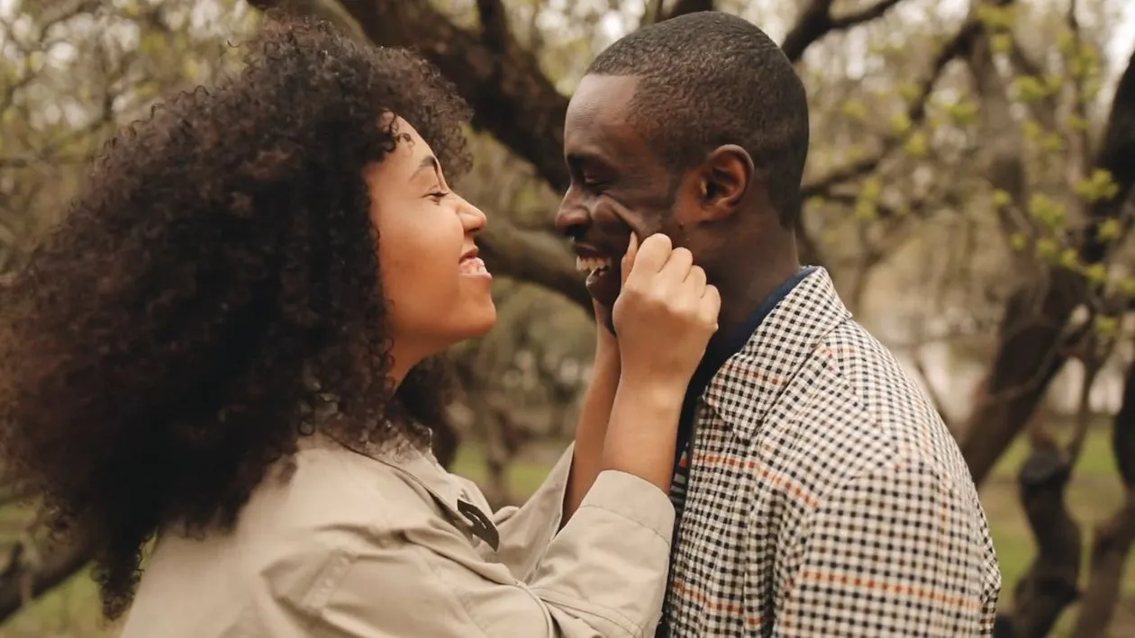 A couple smiling and holding each other’s faces outdoors.